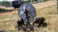 Zwei Soldaten knien auf einer Wiese und arbeiten an einer Satellitenschüssel.
