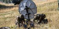 Zwei Soldaten knien auf einer Wiese und arbeiten an einer Satellitenschüssel.
