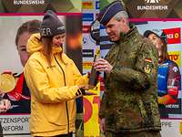 Ein Soldat in Uniform überreicht einer Frau in gelber Jacke und Mütze einen Award.