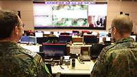 Two soldiers stand in front of monitors in a large room