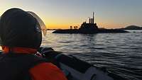 Ein Soldat sitzt in einem Schlauchboot. Im Hintergrund steht ein U‑Boot an der Wasseroberfläche.