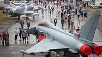 Besucher gehen über ein Gelände auf dem zwei Kampfflugzeuge stehen.