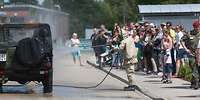 Besucher beobachten eine Person in ABC-Schutzkleidung beim Absprühen eines Militärfahrzeuges.