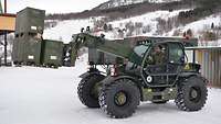 Ein Soldat fährt mit einem Fahrzeug mit Teleskoplader im Gelände und transportiert Materialkisten.