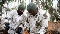 Zwei Soldaten in Schneeflecktarn füllen mit einen Klappspaten einen Sandsack.
