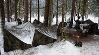 Ein verschneites Zelt steht an einer Feuerstelle. Im Hintergrund packen Soldaten Ausrüstung aus.