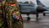 Das Orion-2026-Patch am Ärmel einer deutschen Uniform mit unscharfem Flugzeug im Hintergrund.