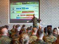 Ein Soldat spricht zu vielen anderen Soldaten und Soldatinnen, im Hintergrund eine Videopräsentation.
