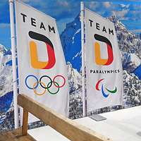 2 Fahnen mit Team D darauf und dem Logo von Olympia und Paralympics, dahinter verschneite Berge