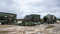 Soldaten beauen ein Zelt auf. Im Hintergrund 3 Lastwagen