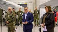Drei Personen vor Mikrofonen vor Presse und weitere Soldaten im Hintergrund