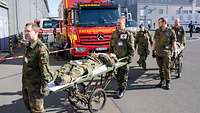 Soldaten transportieren Verletzte auf Rolltragen. Im Hintergrund ein ziviles Rettungsfahrzeug.