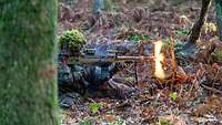 Ein Soldat liegt mit einem MG5 auf Waldboden und schießt. Helles Mündungsfeuer flammt auf.