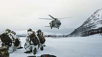 Gebirgsjäger knien im Schnee, ein Transporthubschrauber landet im Hintergrund.
