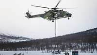 Ein Transporthubschrauber setzt Außenlast im Schnee ab, Soldaten sichern am Boden.