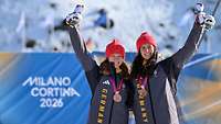 Zwei Ski-Fahrerinnen jubeln bei der Medaillenzeremonie und halten ihre Maskottchen hoch.