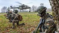 Mehrere bewaffnete Soldaten knien auf einer Wiese während ein Hubschrauber landet.