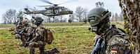 Mehrere bewaffnete Soldaten knien auf einer Wiese während ein Hubschrauber landet.