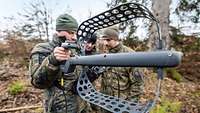 Ein Soldat im Gelände hält ein Gerät in Hand. Zwei Soldaten stehen daneben.