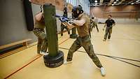 Soldaten mit Kopfschutz beim Nahkampf mit blauen Übungsgewehren und Sandsäcken in einer Turnhalle.