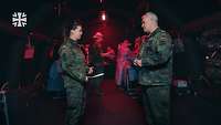 Eine Soldatin und ein Soldat stehen sich gegenüber und sprechen miteinander. Im Hintergrund sind Personen in OP-Kleidung.