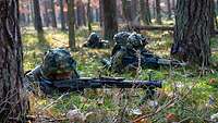 Drei Soldaten liegen zwischen Bäumen mit Gewehr im Anschlag auf grünem Waldboden.