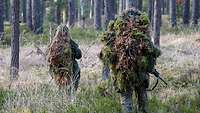 Drei mit Moos getarnte Soldaten gehen mit Gewehren durch den Wald.
