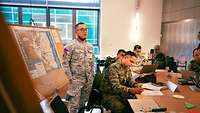 Menschen mit Uniform sitzen an Tischen, ein Soldat steht neben einer Landkarte.