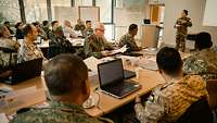 Eine Soldatin steht vor Kursteilnehmern, die an Tischen sitzen und zuhören.