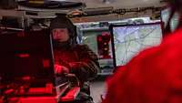 Ein Soldat sitzt vor einem Gerät in einem rot beleuchtet Innenraum eines Gefechtsfahrzeuges.