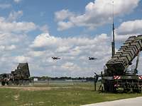 Zwei Tornado-Kampfjets fliegen in Formation über Patriotsysteme, die am Boden stehen.