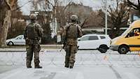 Zwei Soldaten stehen hinter einem am Boden liegenden Stacheldraht. Autos fahren vorbei.