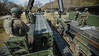 Soldaten stehen auf zwei Fahrzeugen und transportieren Brückensegmente mit einen Kran.