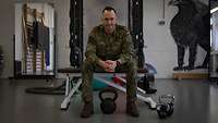Ein Soldat sitzt auf einer Hantelbank, im Hintergrund sind Sportgeräte zu sehen.