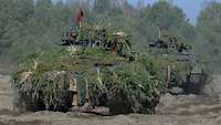 Zwei mit Zweigen getarnte Panzer fahren hintereinander im Gelände.