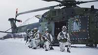 Soldaten mit Waffen knien vor einem Hubschrauber in einer Schneelandschaft.