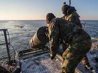Zwei Soldaten auf hoher See schieben eine Metalltonne von Bord eines Schiffes ins Wasser.