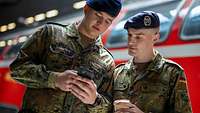 Zwei Soldaten stehen nebeneinander und schauen auf ein Mobiltelefon. Im Hintergrund steht ein Zug.