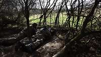 Zwei Soldaten liegen in einer Stellung am Waldrand und schauen auf das Gelände vor ihnen..