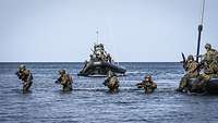 Vier Soldaten stehen im Wasser mit ihren Waffen im Anschlag und zwei Schlauchboote mit Soldaten.