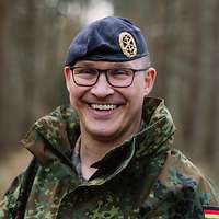 Ein Soldat mit blauem Barett steht im Wald und lächelt in die Kamera.