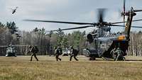 Soldaten mit Ausrüstung verlassen einen Hubschrauber und laufen über eine Wiese.