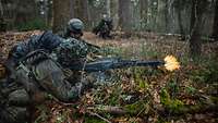 Ein Soldat liegt im Wald auf dem Boden und schießt. Neben ihm liegt ein anderer Soldat.