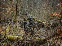 Ein Soldat kniet mit seiner Waffe zwischen Ästen und Büschen im Unterholz.