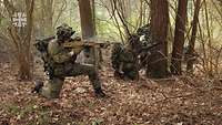 Ein Soldat kniet mit seiner Waffe im Anschlag im Wald. Neben ihm anderen Soldaten in Stellung.