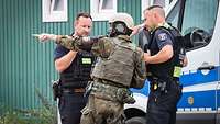 Ein Soldat mit Schutzweste und Helm weist zwei Polizisten vor einem Polizeifahrzeug in eine Lage ein.