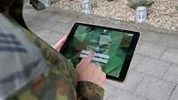 Über die Schulter eines Soldaten blickt man auf ein Tablet. Darauf ist die App und der Schriftzug USG-Online zu sehen.