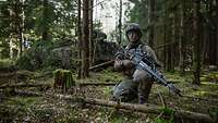 Ein Soldat mit Waffe kniet im Wald. Im Hintergrund steht ein mit Zweigen getarnter Panzer.