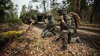 Zwei Soldaten knien im Gelände vor einem Panzer. Einer von beiden spricht in ein Funkgerät.