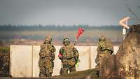 Drei Soldaten stehen hinter einer Mauer, einer davon hat eine rote Flagge in der Hand.